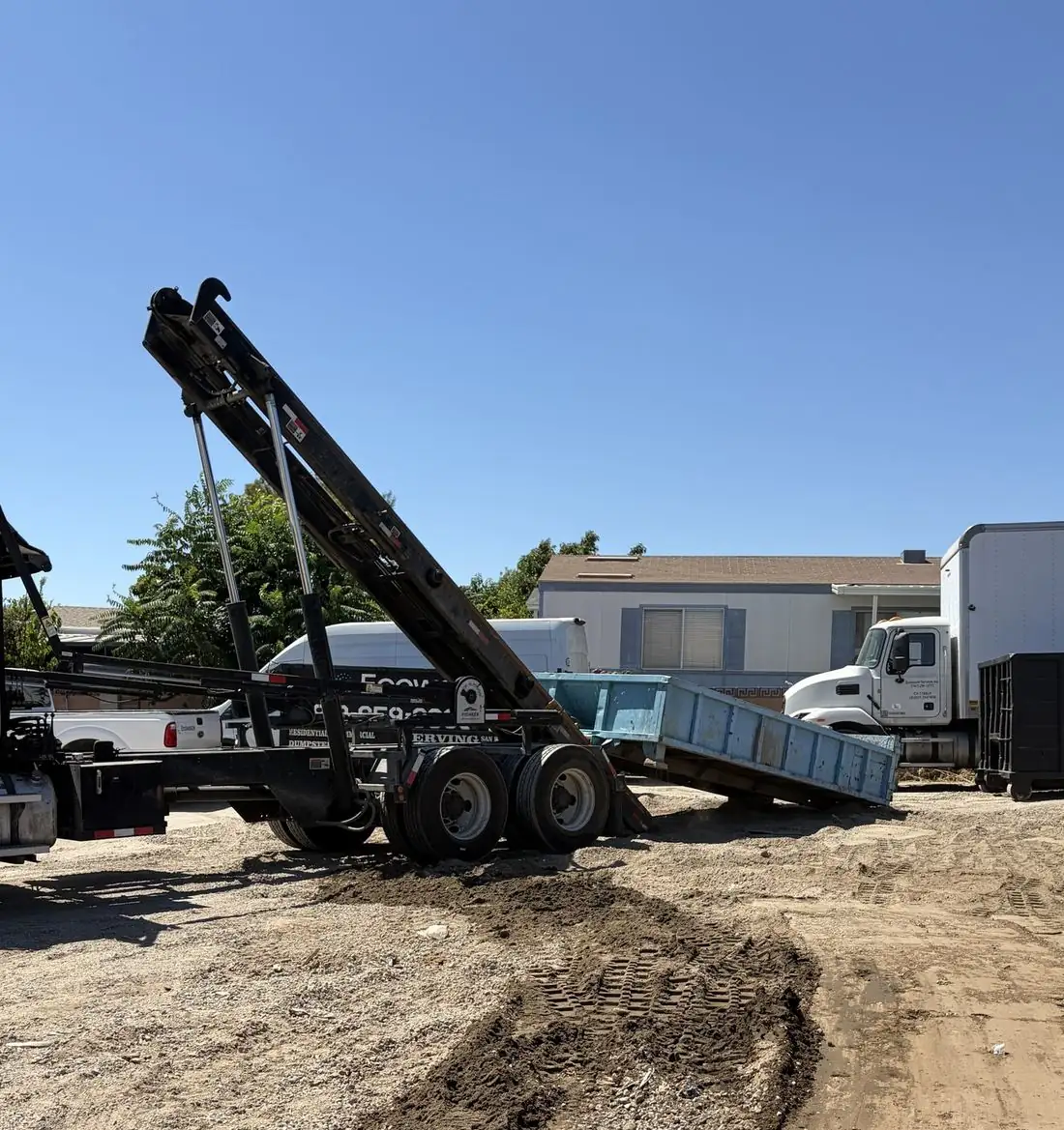 Roll-off truck lifting a blue roll-off dumpster onto its bed at a dirt job site